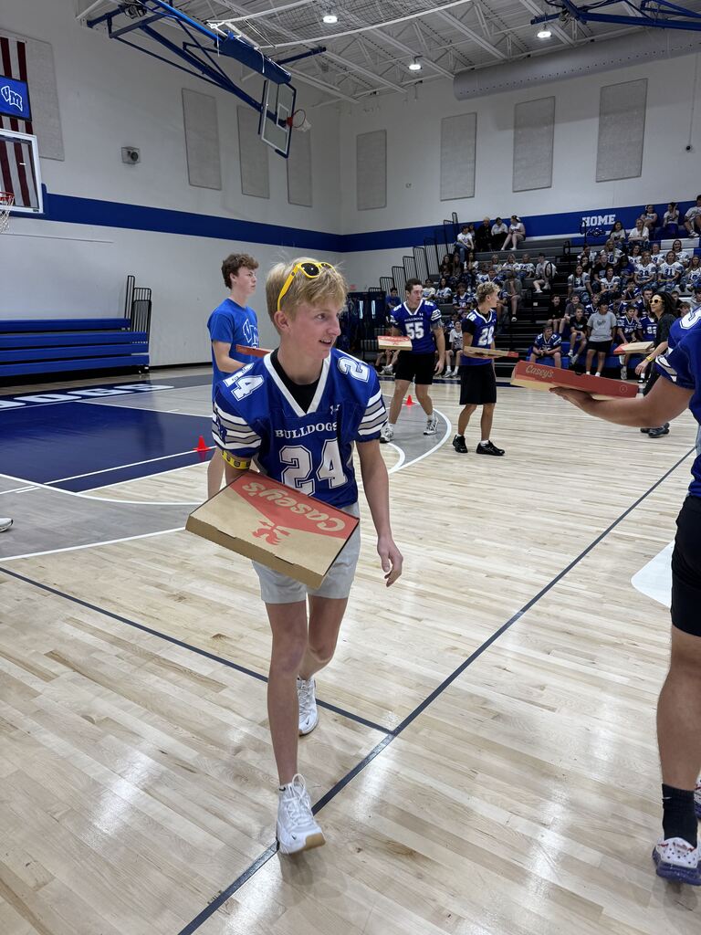 Student in a blue Bulldogs football jersey carries a pizza box across the gym during a school event.