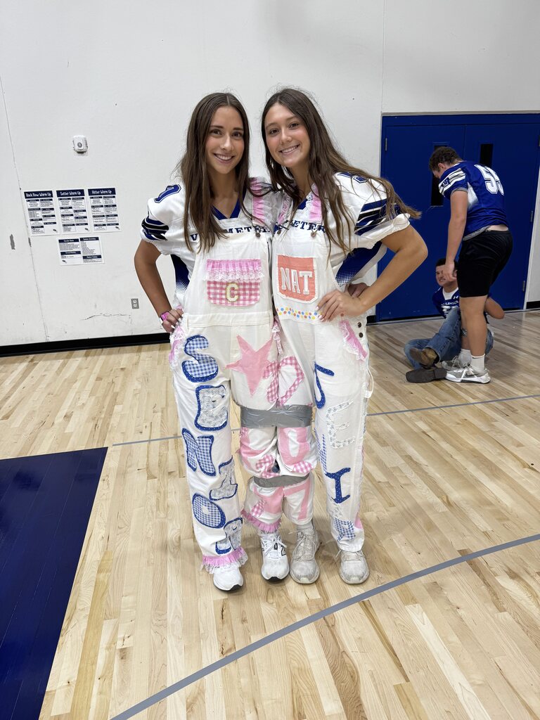 Two students in white football jerseys pose together in matching decorated overalls with pink and blue details.