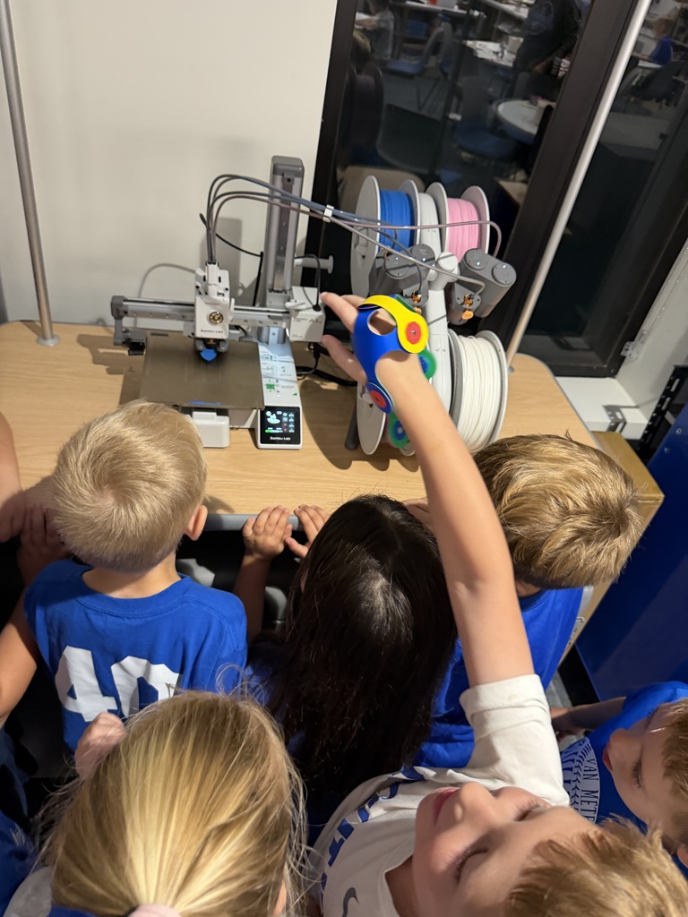 Children gathered around a 3D printer observing the printing process with colorful filament spools in the background.
