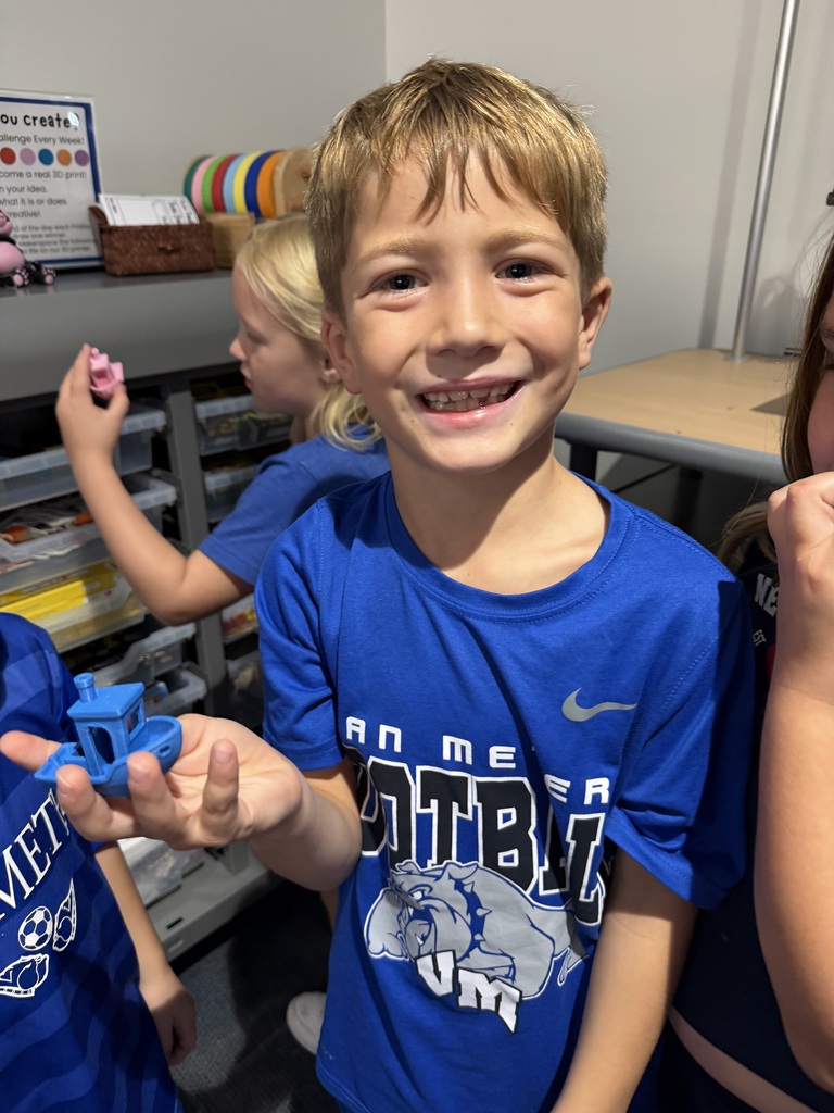 Smiling boy in a blue football t-shirt holding a small blue toy in a classroom setting.