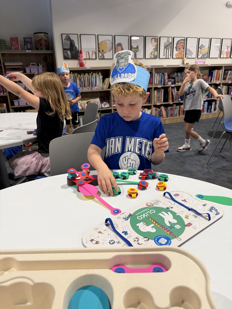 Young boy wearing a blue Bulldogs paper crown plays with colorful fidget spinners at a library table filled with books and educational materials.