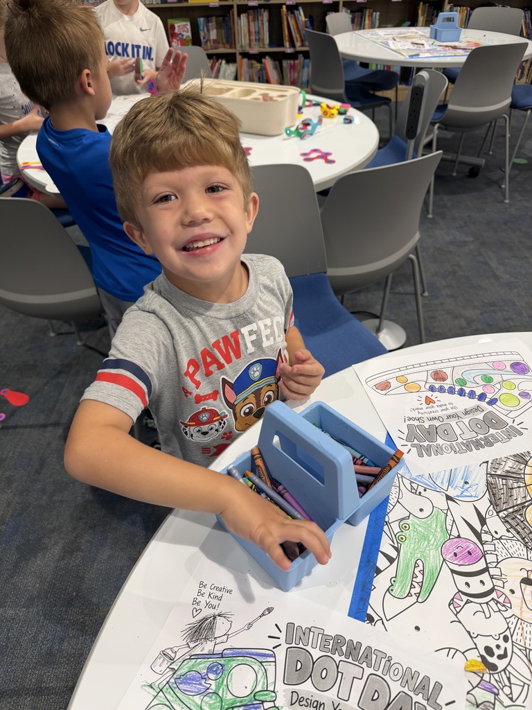 Young boy smiling while coloring a detailed International Dot Day poster at a classroom table filled with crayons and art supplies.