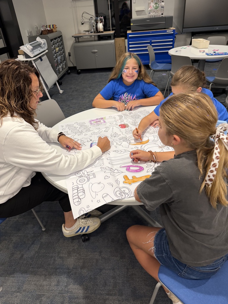 Three children and an adult woman coloring a large poster filled with outlined drawings at a round table in a classroom setting.