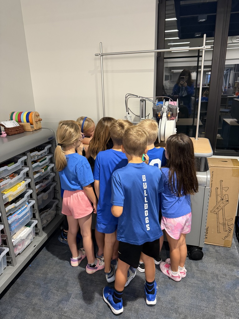 A group of young children in blue shirts closely gathered around a 3D printer in a classroom setting.