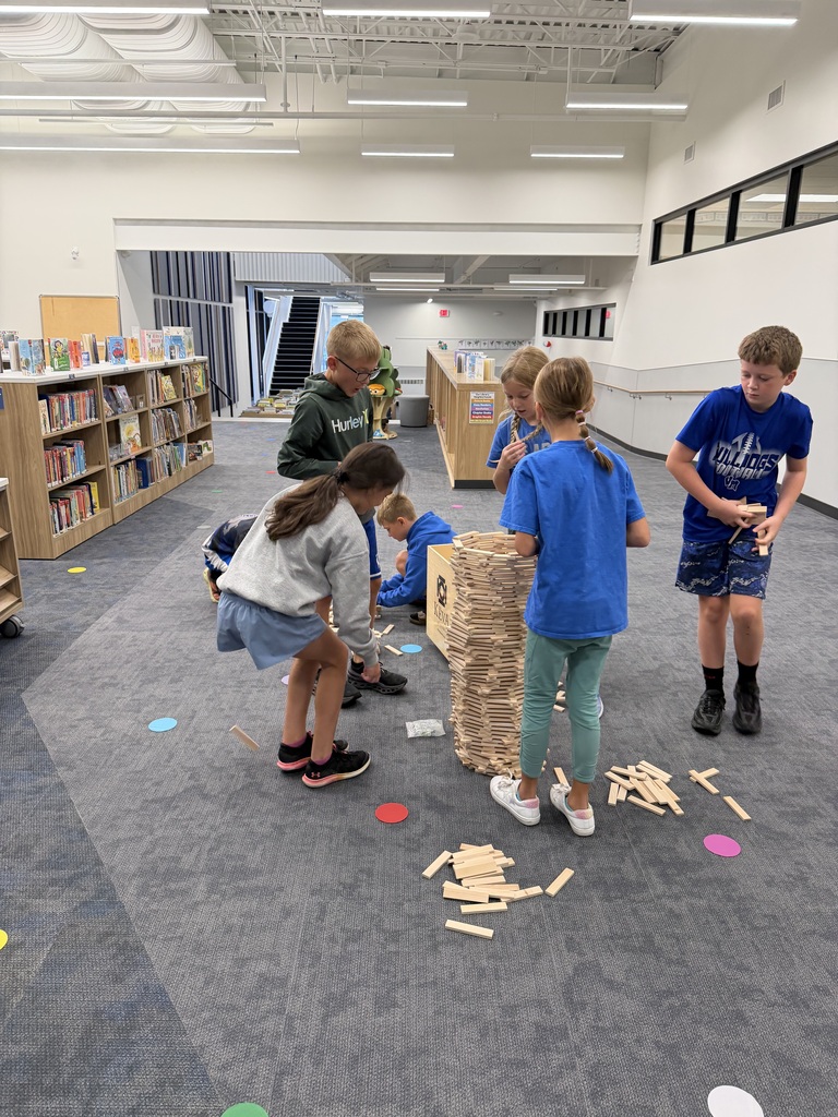 Children building a tall tower with wooden blocks in a spacious library area surrounded by bookshelves.