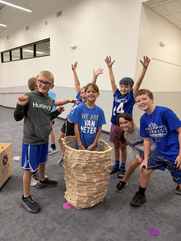 Group of seven kids smiling and posing indoors, one standing inside a tall structure made of wooden blocks.