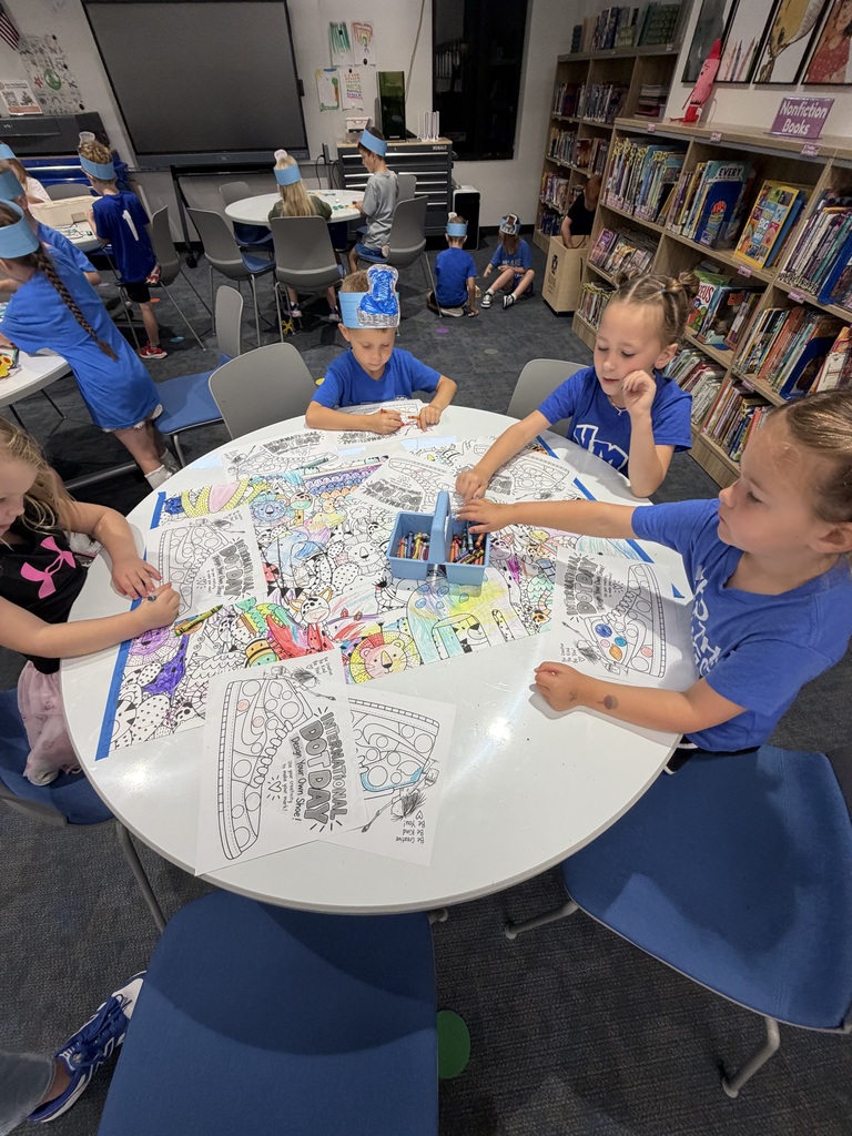 Children seated around a round table coloring detailed activity sheets in a library setting with bookshelves in the background.