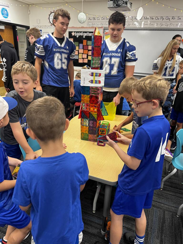 Younger students build around a tall magnetic-tile tower while older students in Bulldogs jerseys assist nearby.