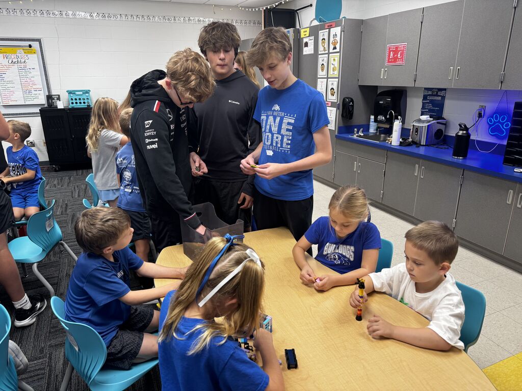Older and younger students gather around a classroom table, building and examining small colorful construction pieces.