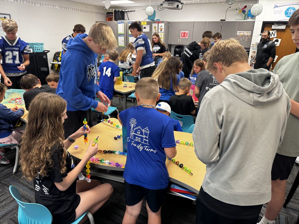A busy classroom of older and younger students collaborates on colorful interlocking chains at several tables.