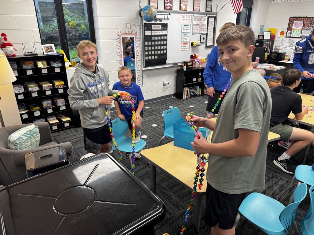 Students smile while connecting colorful flower-shaped pieces into long chains during a collaborative classroom activity.