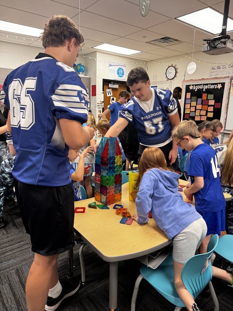 Older students in blue Bulldogs jerseys help younger students build a tall, colorful structure with magnetic tiles.