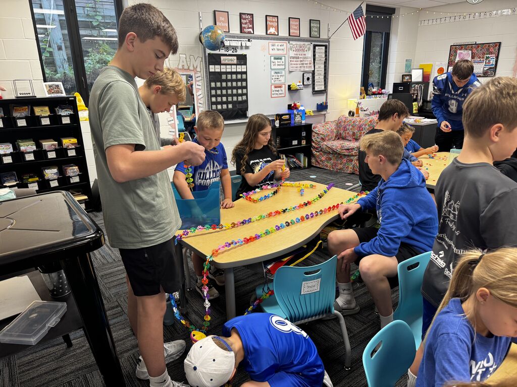 Students work around a table to assemble long, multicolored chains from small interlocking flower-shaped pieces.