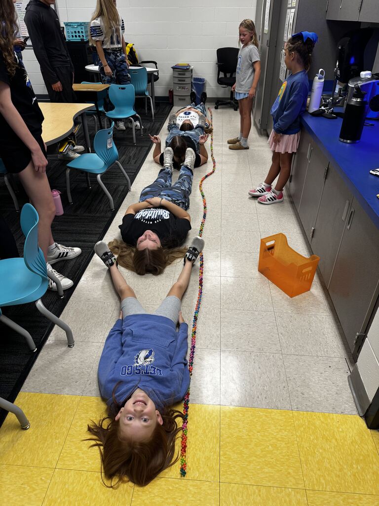 Students lie head-to-toe on the classroom floor beside a long multicolored chain, appearing to compare their combined length.