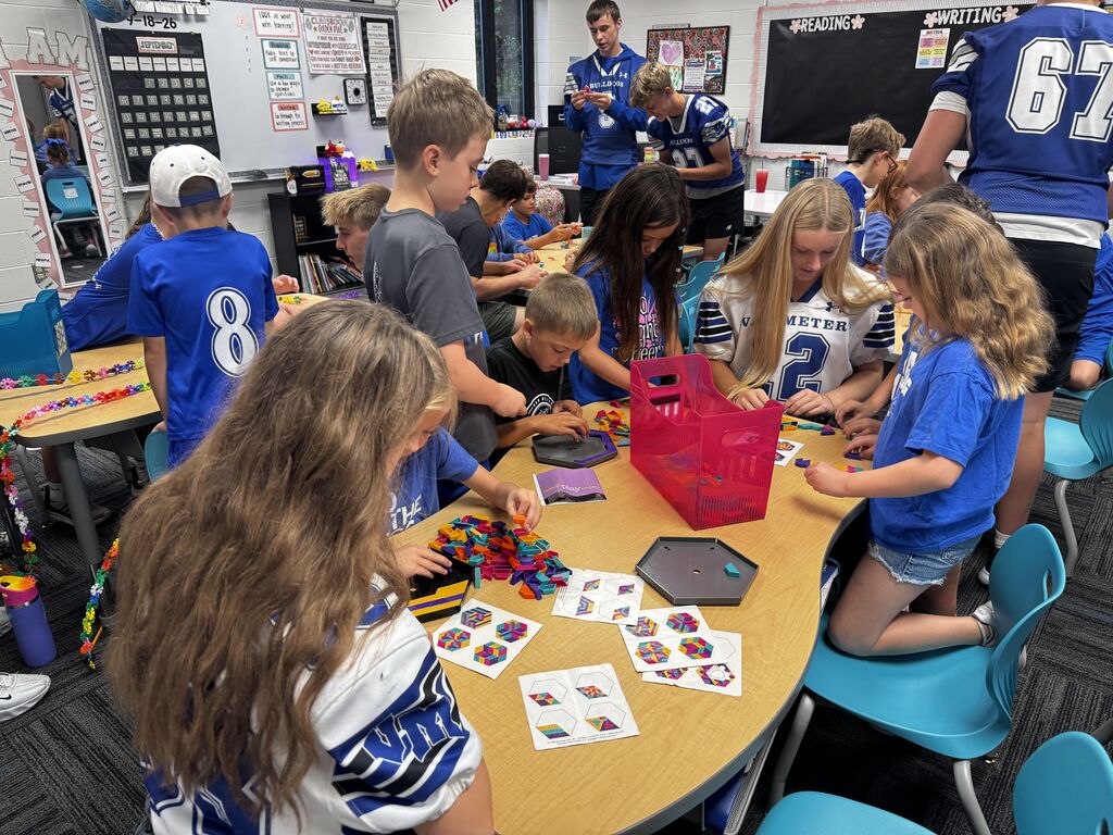 Students work together around a classroom table, arranging bright geometric tiles into colorful patterns and designs.