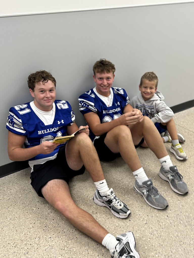 Two older football players and a young boy smile while sitting together against a school hallway wall.