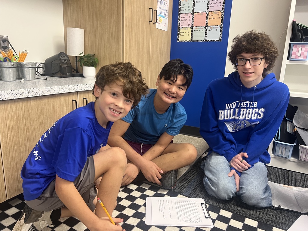 Three students sit together on a checkered classroom rug with a clipboard and worksheet in front of them.