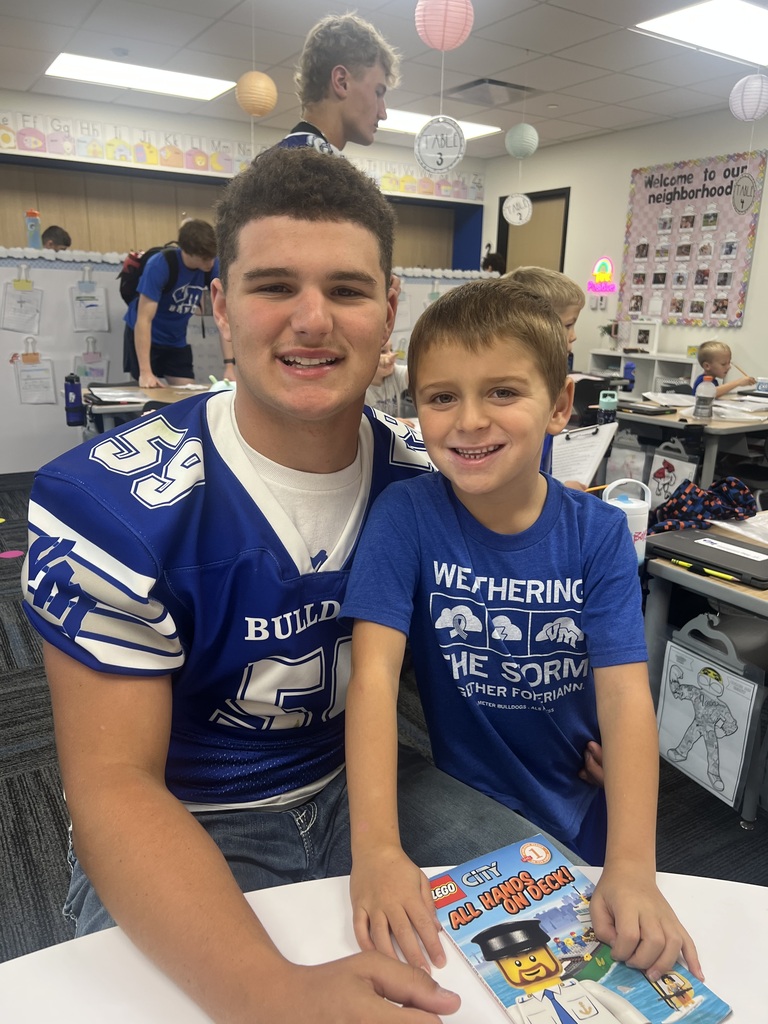 An older student in a blue football jersey and a smiling young boy pose together with a LEGO book.