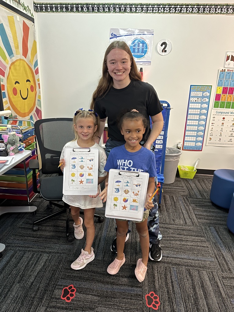 Two young girls proudly hold completed worksheets while an older student smiles behind them.