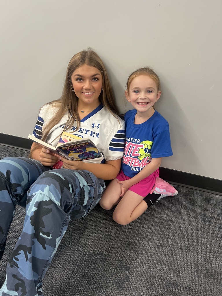 An older student and a smiling young girl sit together on the floor with an open chapter book.