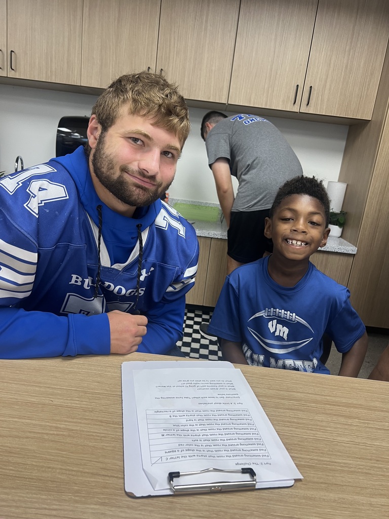 An older football player and a smiling young boy pose together at a classroom table with a worksheet.
