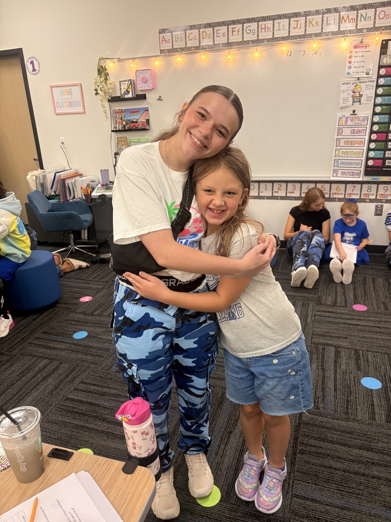 An older student and a young girl smile and hug in a classroom while other students work in the background.