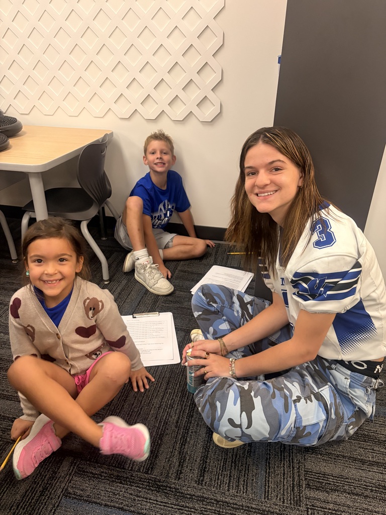 An older student and two smiling children sit on the classroom floor with worksheets and pencils nearby.