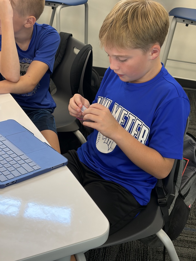 Young boy in a blue T-shirt focused on assembling a small object while sitting at a desk with a laptop nearby.