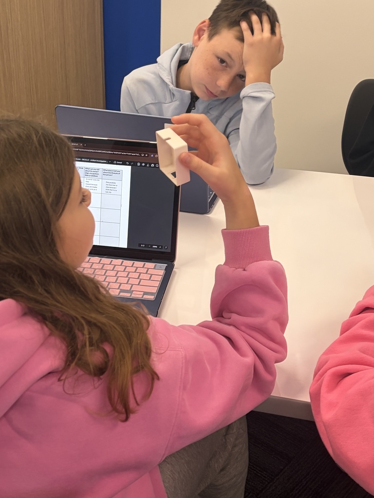 Girl in pink sweatshirt examining a small white object while sitting at a table with a laptop, boy in background looking thoughtful with hand on forehead.