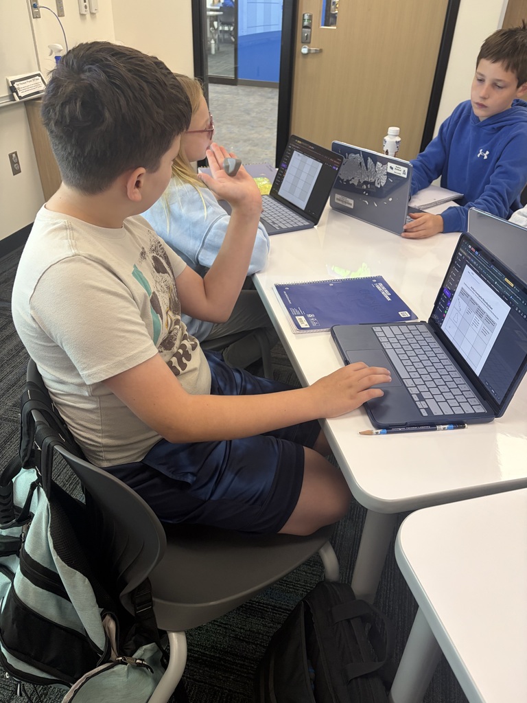 Three students sitting at a table working on laptops and discussing a project in a classroom setting.