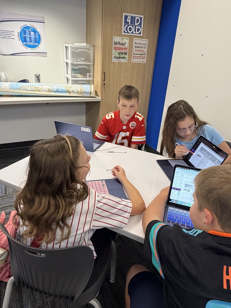 Four children sitting around a white table working on laptops in a classroom setting with educational posters on the wall.