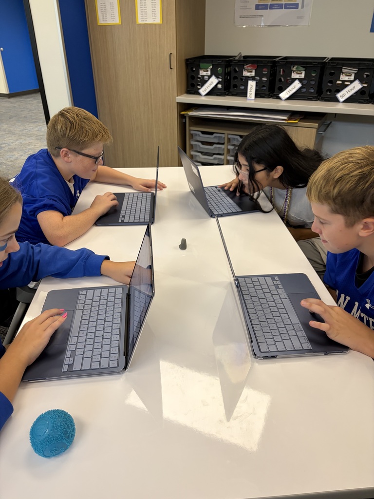 Four students seated around a white table, each focused on their laptops in a classroom setting.