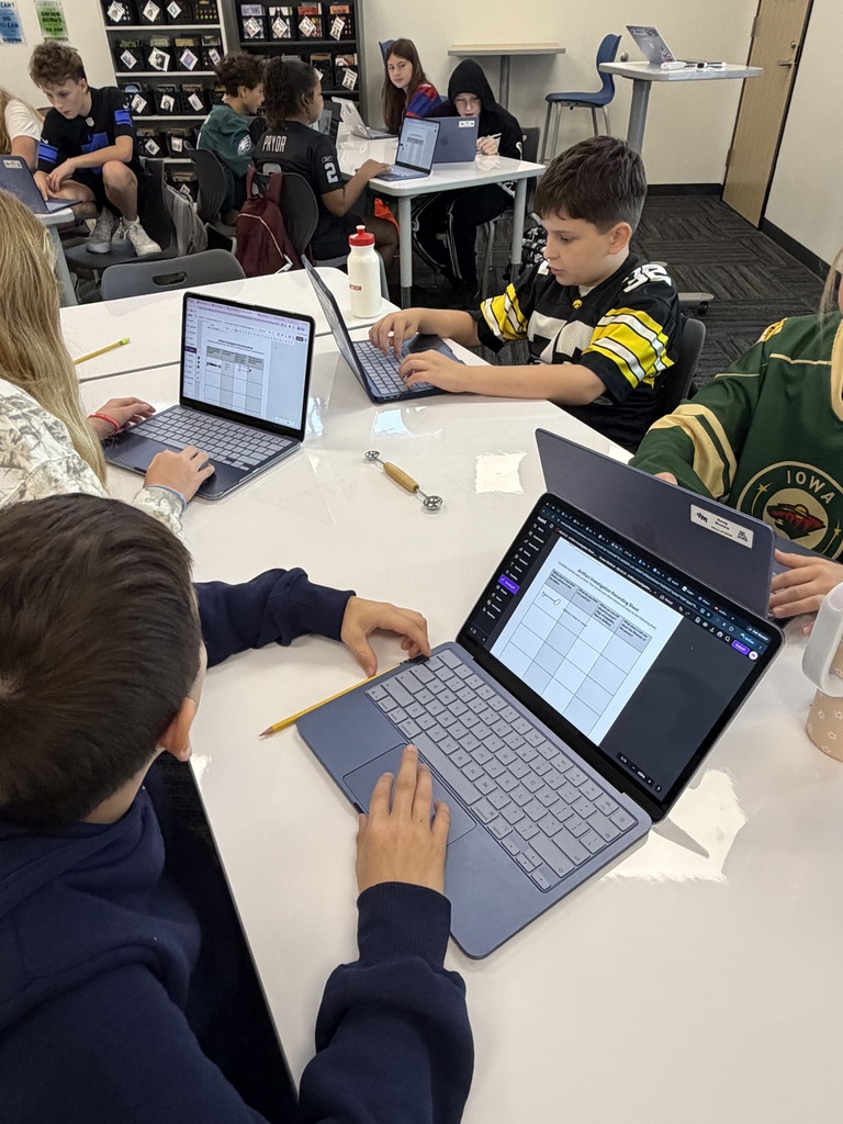 Students working on laptops around a classroom table, focused on digital spreadsheets and assignments.