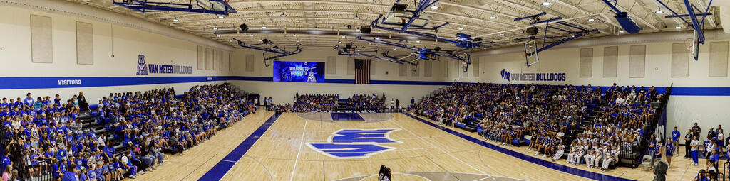Panoramic view of a high school gym filled with students seated on bleachers during a school event, with a large blue logo on the basketball court floor.