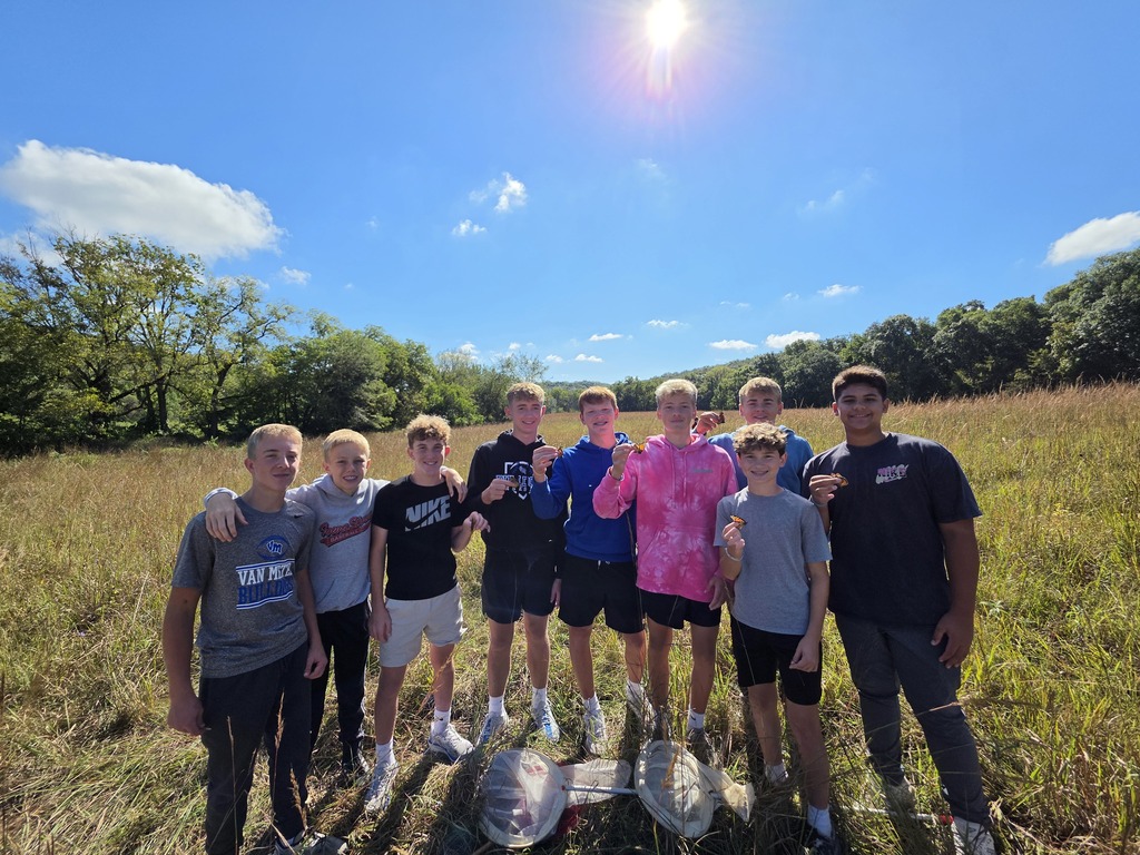 Nine teens pose together in a sunny prairie, several holding monarch butterflies after collecting them in the field.