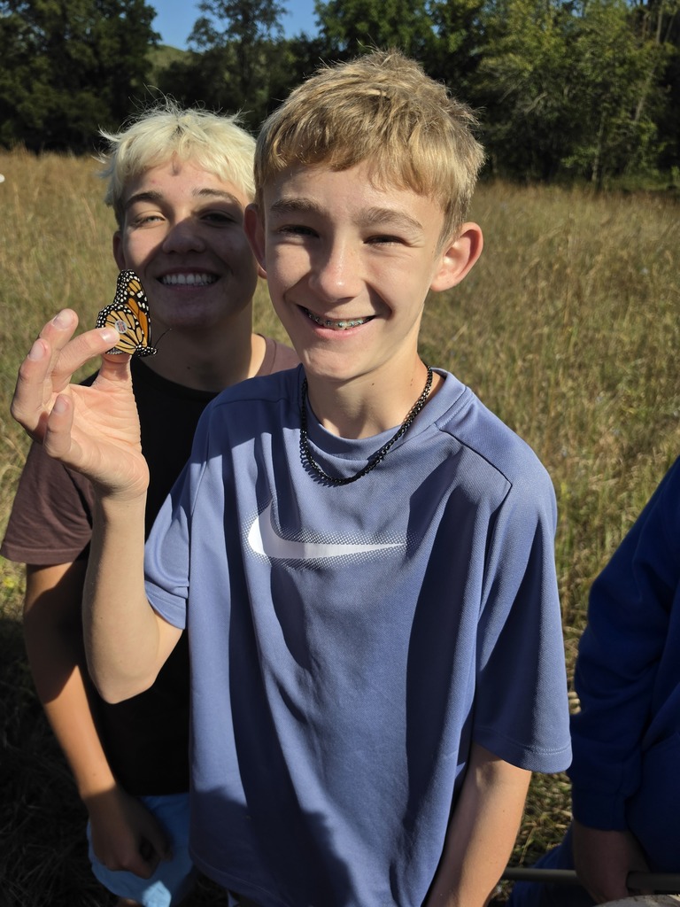 Smiling teen holds a monarch butterfly on his fingers while another teen smiles behind him in a sunny prairie.