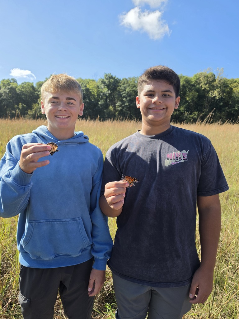 Two smiling teens each hold a monarch butterfly while standing together in a sunlit grassy prairie.