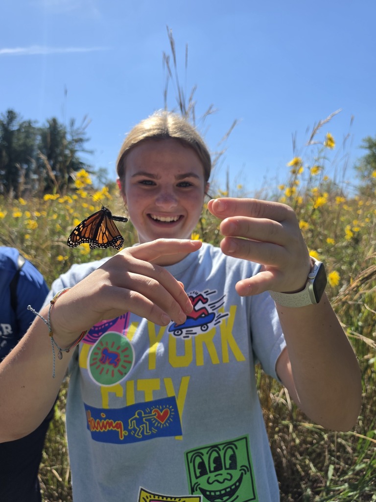 Smiling student reaches toward a monarch butterfly perched on her hand amid yellow wildflowers and tall prairie grass.