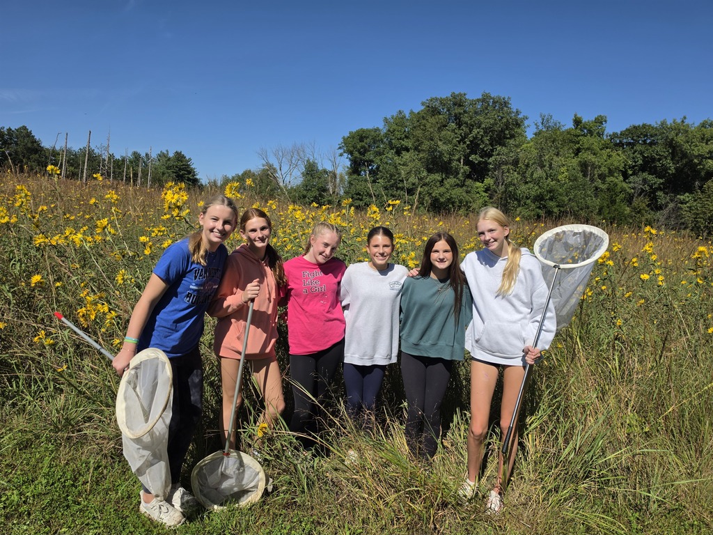 Six students with butterfly nets pose among tall grasses and yellow wildflowers under a bright blue sky.
