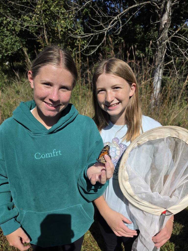 Two smiling students pose outdoors with a monarch butterfly perched on one student’s hand beside a butterfly net.