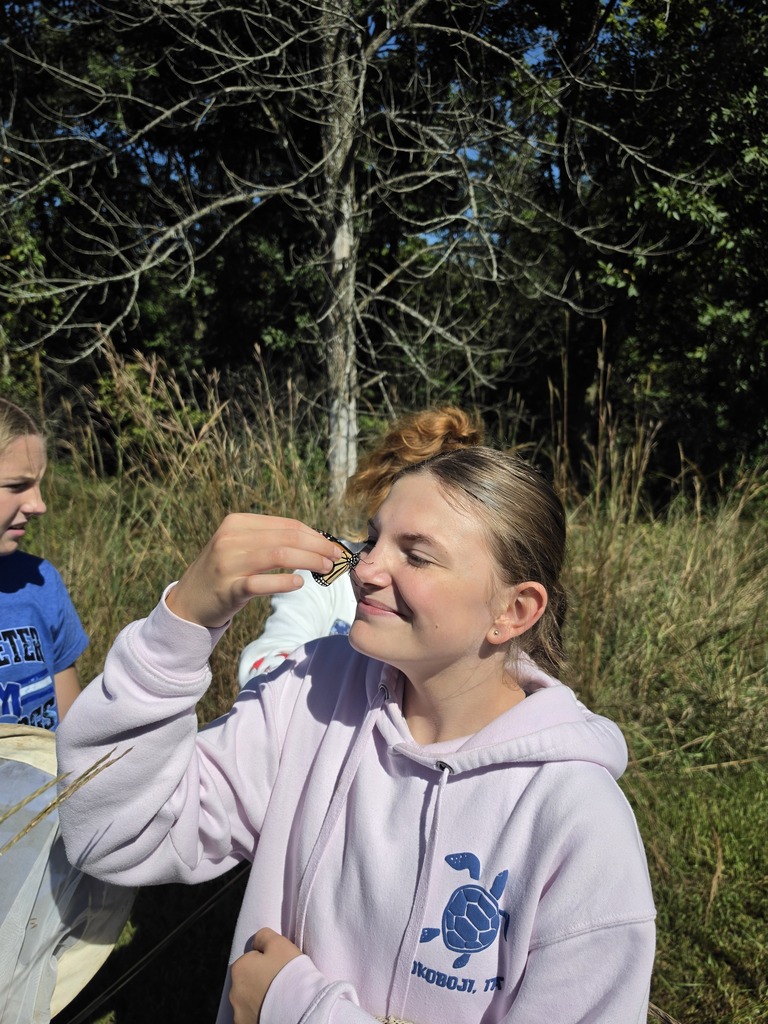 Smiling student in a pale pink hoodie holds a monarch butterfly near her face in a sunny grassy field.