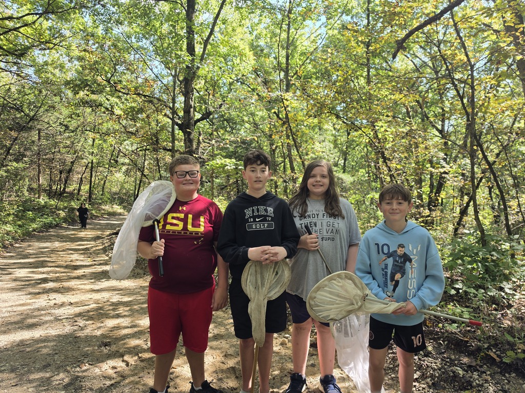 Four students stand on a wooded trail holding large butterfly nets during an outdoor field activity.