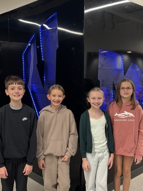 Four smiling children standing indoors against a black wall with blue LED lights.