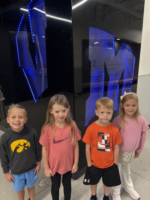 Four smiling children standing indoors in front of a black wall with large blue illuminated letters.
