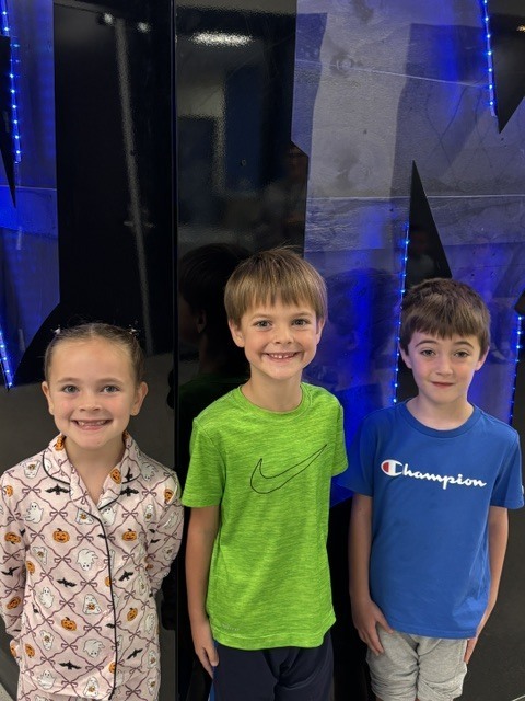 Three smiling children stand side by side against a dark backdrop with blue lights, wearing casual colorful shirts.