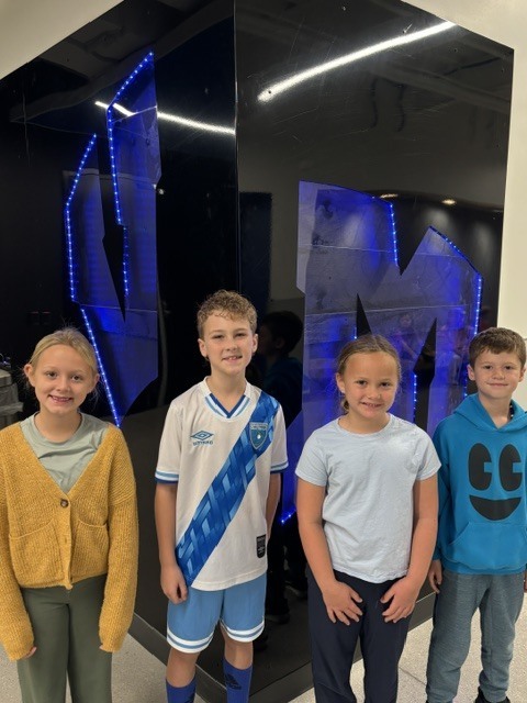 Four smiling children stand in front of a black wall with blue illuminated geometric shapes indoors.