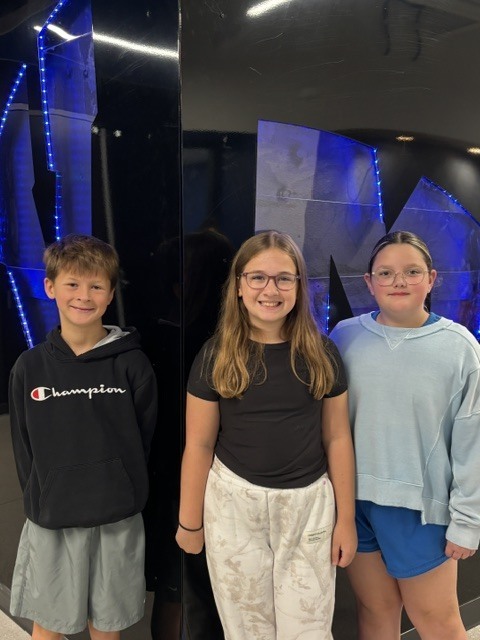 Three smiling children stand indoors against a dark wall with blue LED lights, dressed casually in hoodies and shorts.