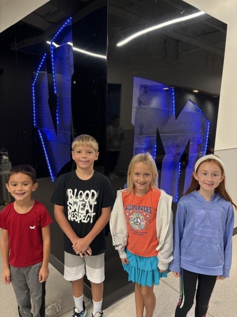 Four smiling children stand indoors in front of a black wall with a large illuminated blue letters V and M.