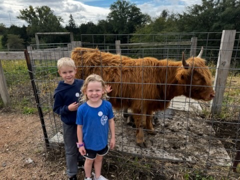 Two smiling children pose beside a shaggy brown Highland cow behind a wire fence on a cloudy day.
