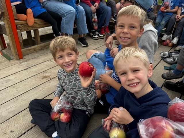 Three smiling boys sit together on a wooden floor, holding bags of freshly picked apples; one proudly displays a red apple.